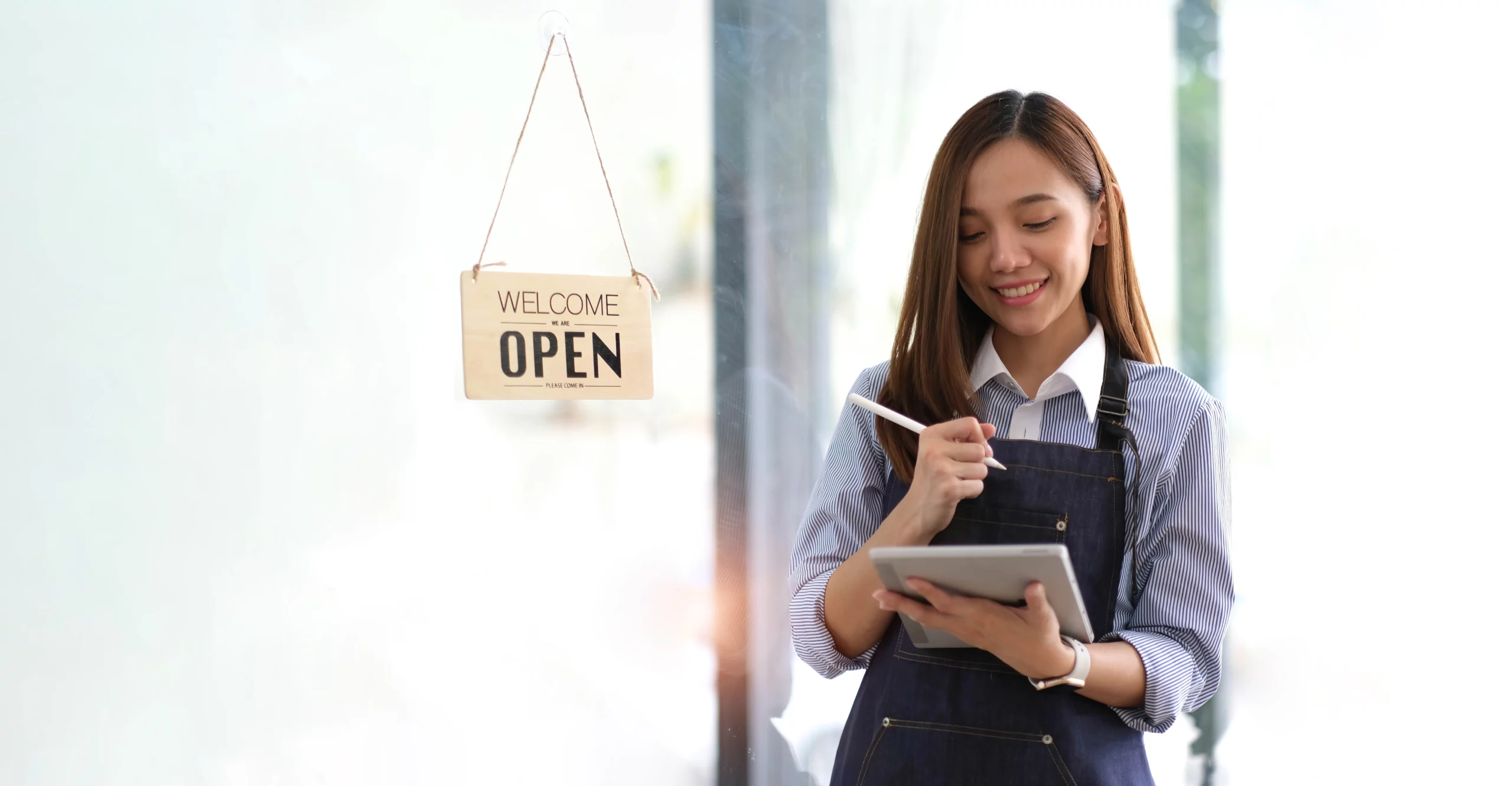 Smiling small business owner in apron checking tablet beside ‘Welcome Open’ sign — representing business license compliance in Florida