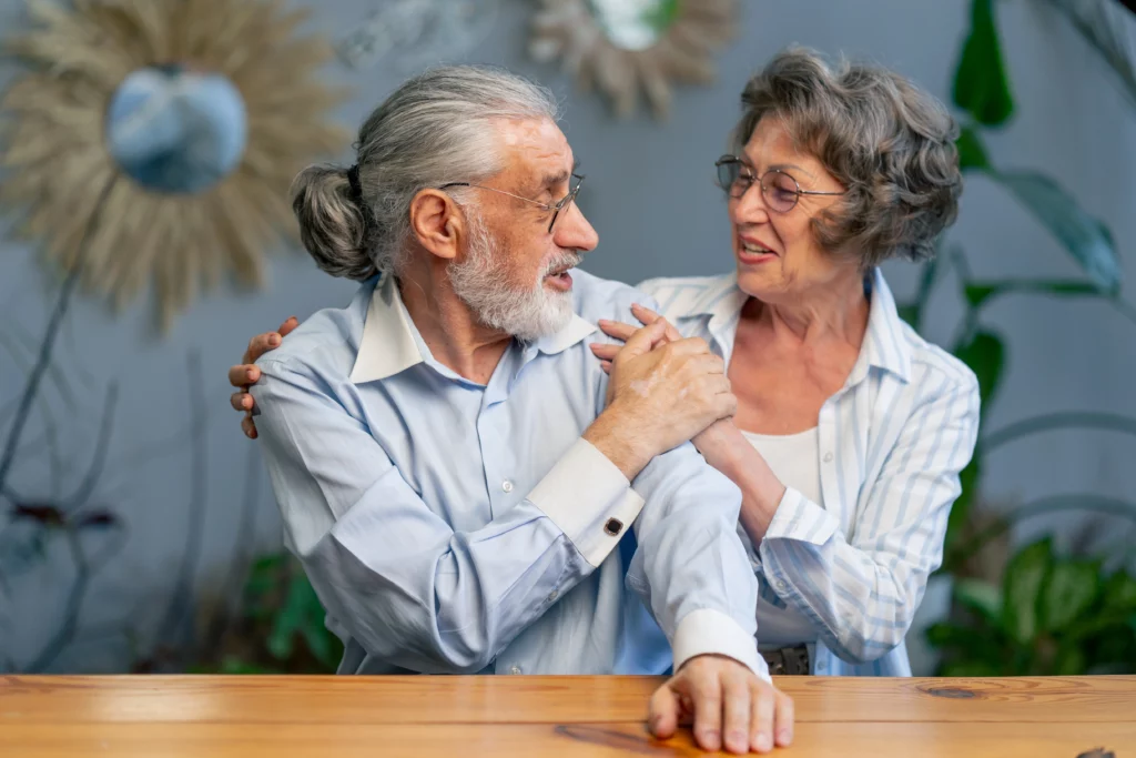 Elderly couple smiling at each other in cozy setting — woman’s hand on man’s shoulder in affectionate moment