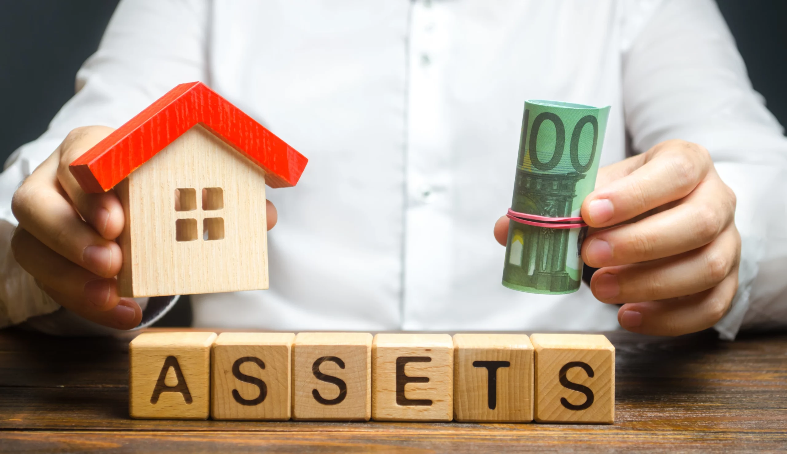Person holding wooden house model and roll of euro bills above wooden blocks spelling “ASSETS” — representing probate vs non-probate assets