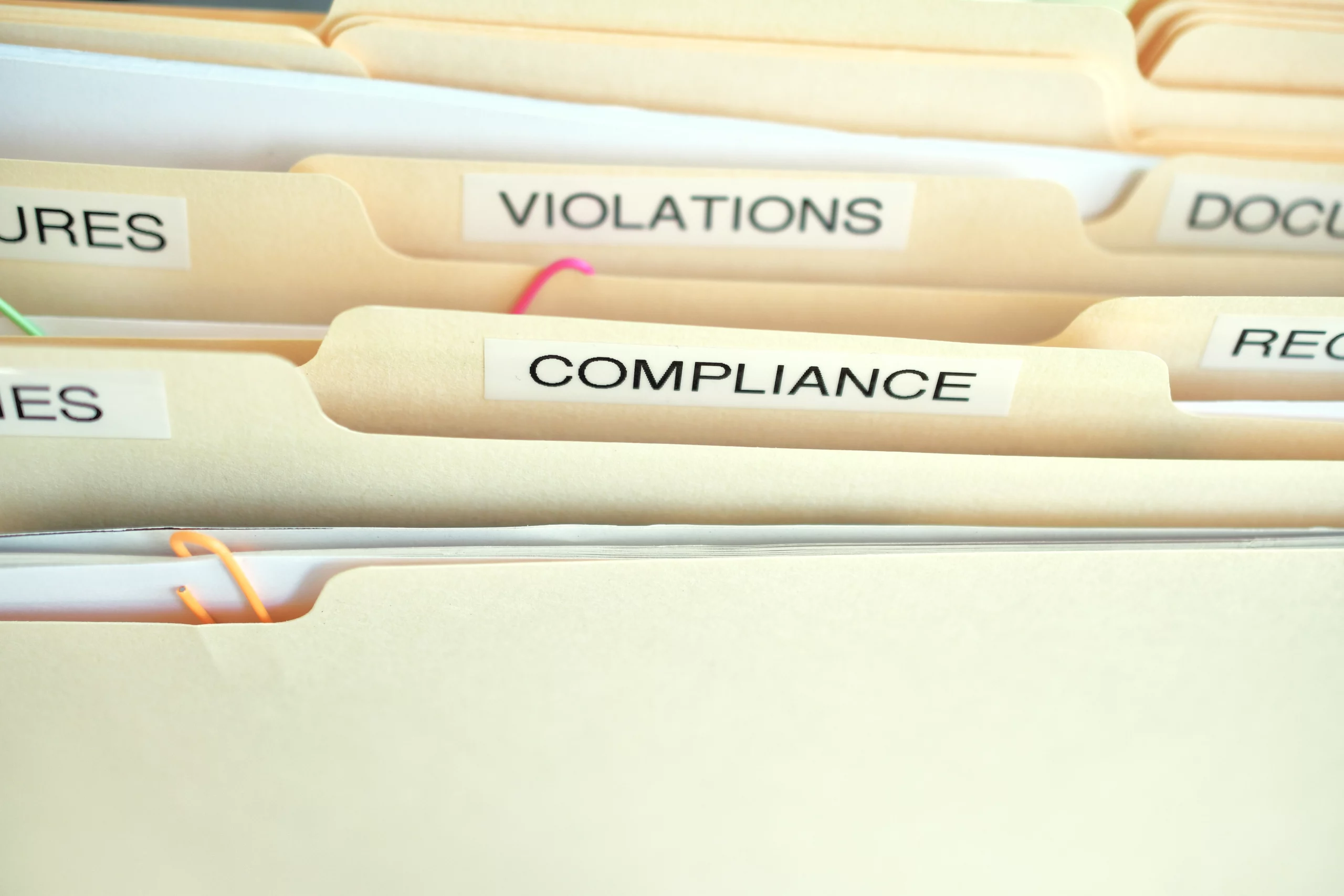 Close‑up of neatly organized beige filing folders labeled “COMPLIANCE,” “VIOLATIONS,” and “PROCEDURES” — symbolizing structured record‑keeping, regulatory adherence and employment law compliance for small businesses.