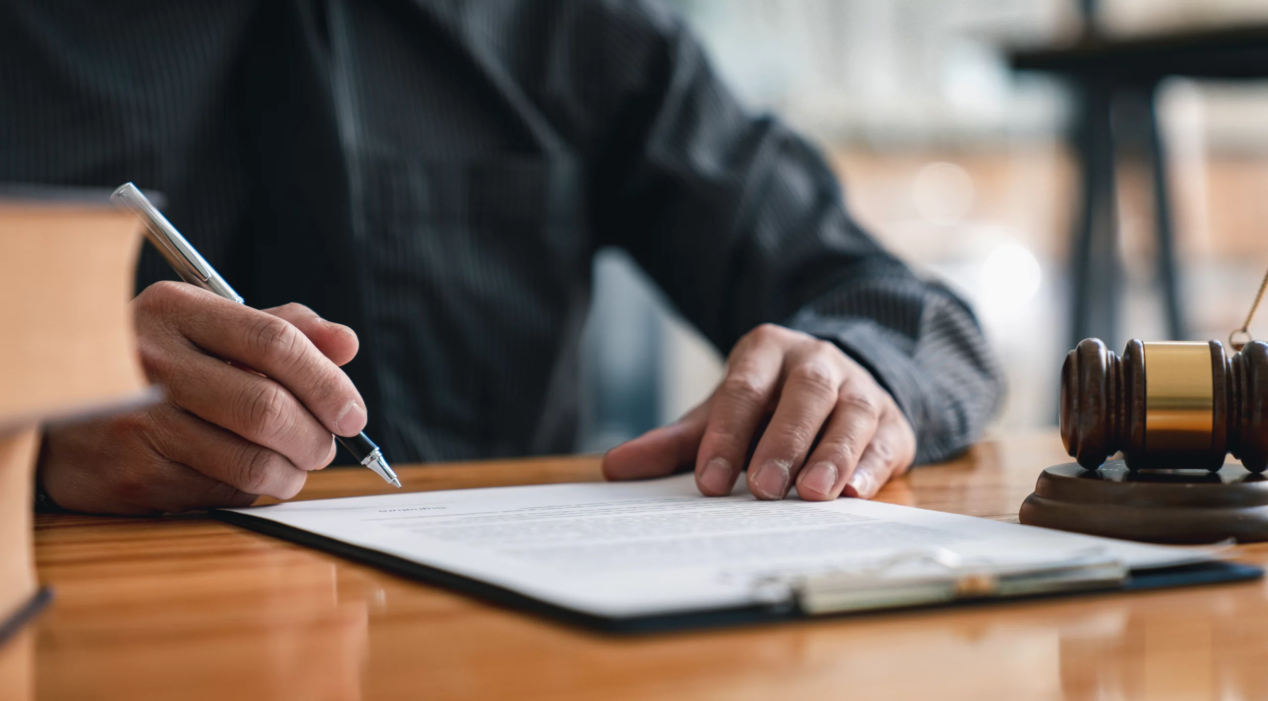 Person in a dark shirt signing a legal document at a wooden desk, with a gavel and law books nearby — representing formal legal paperwork, power of attorney execution and estate planning.
