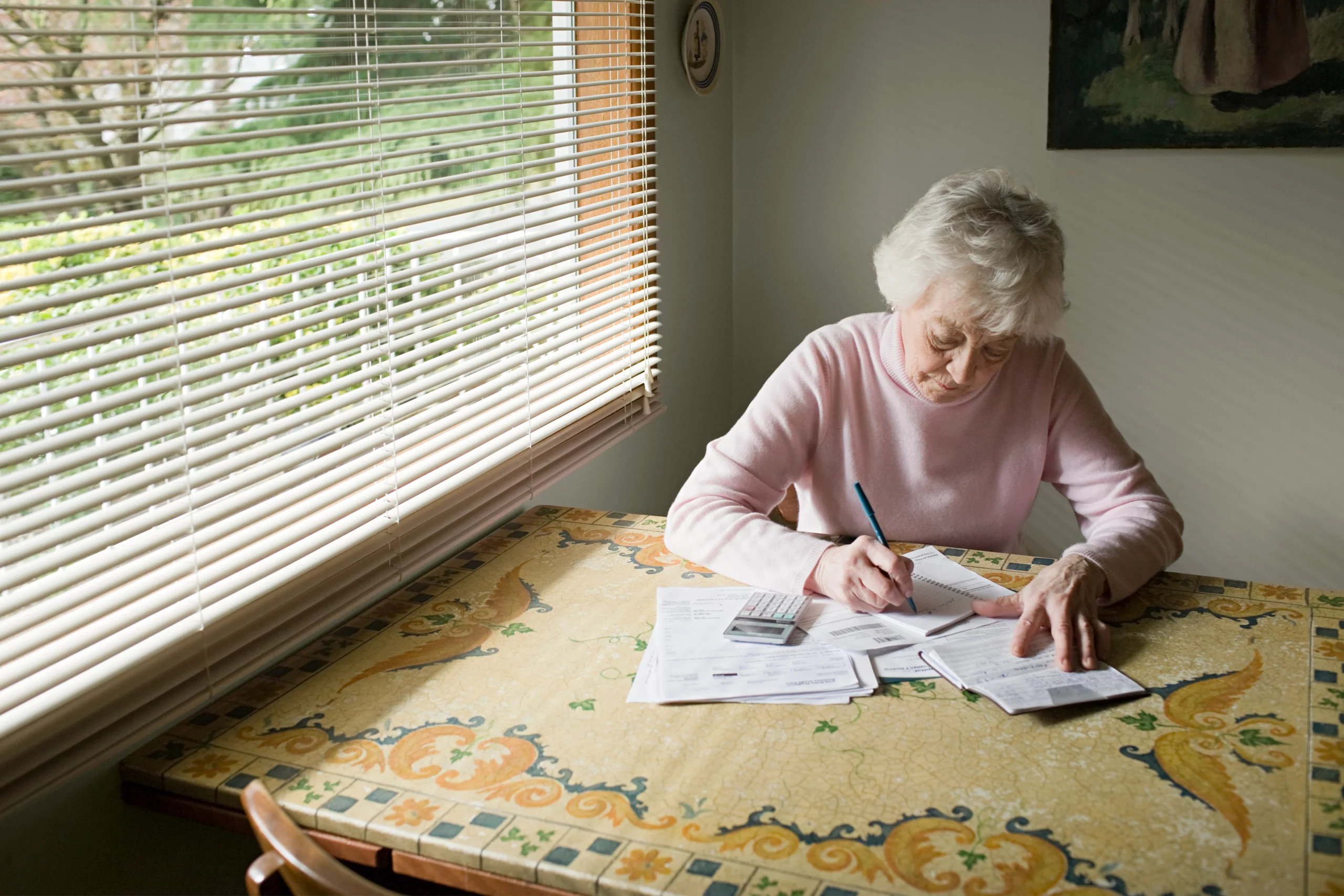 Elderly woman writing on paperwork at a table in sunlight, illustrating preparing documents for a quit claim deed property transfer in Florida.